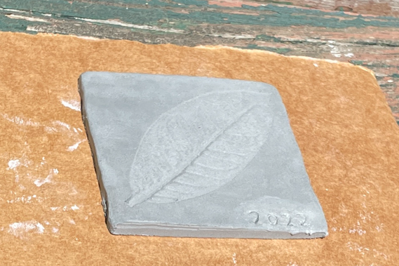 A photo of an grey clay tile resting in the sun on a piece of cardboard. It is unfired clay, set outside to dry. The tile is of an impression of a round leaf. There is a 