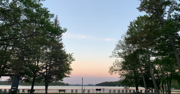A photo of the sunset as seen from the front lawns of CNIB, as looking at the lake. There is white fence between the camera and the lake, with many benches and white muskoka chairs. The sky is blue, peach, pink and purple. There are leafy trees in view.