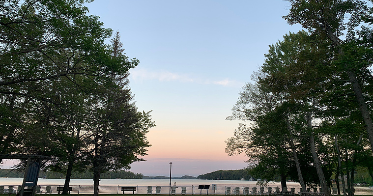 A photo of the sunset as seen from the front lawns of CNIB, as looking at the lake. There is white fence between the camera and the lake, with many benches and white muskoka chairs. The sky is blue, peach, pink and purple. There are leafy trees in view.