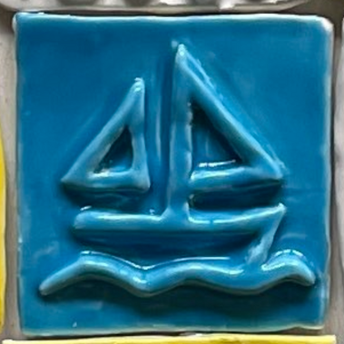 A detail shot of one tile. It is blue and the design is of the Lake Joe sailboat logo, in relief so that the design is raised off the tile and smooth. It is a simple sailboat with a wave underneath.
