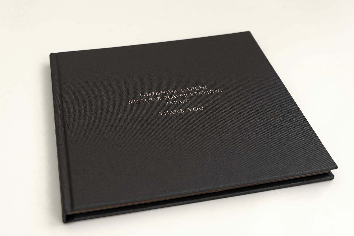 A hardback book, 50cm by 50cm. It is covered in dark grey fabric. Silver text is embossed on the front. It reads 