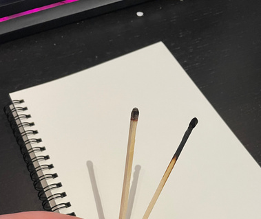 A hand holding two used match sticks over a white sketch pad.