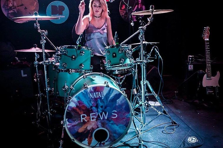 A woman sat behind a teal drumkit on a stage in a small music venue. The bass drum skin uses the same powder paint explosion image but has the band name 