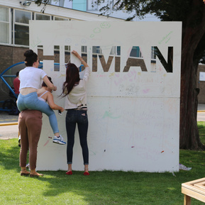 A lady stands in front of the installation and is writing on it. Another person has jumped onto another person's back to reach higher on the installation. 
