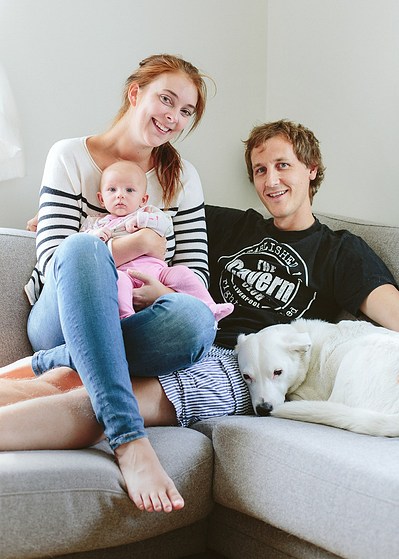 Familie på tre med baby sitter i en sofa med en hvit hund liggende ved siden av