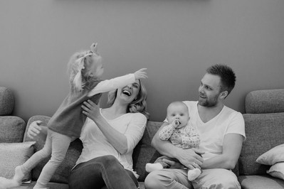 Familie på fire sittende i sofa. Liten jente hopper og kaster seg mot sin mor.