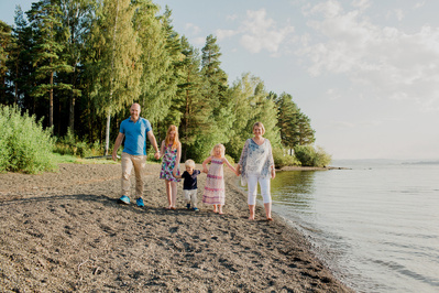 Familie på fem holder hender og går på rekke langs vannkanten en solrik sommerdag