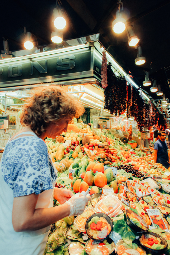 Portrait-oriented up close color photo of shopkeeper marking assorted fresh fruits and vegetables at Barcelona's Las Ramblas famous in-door market