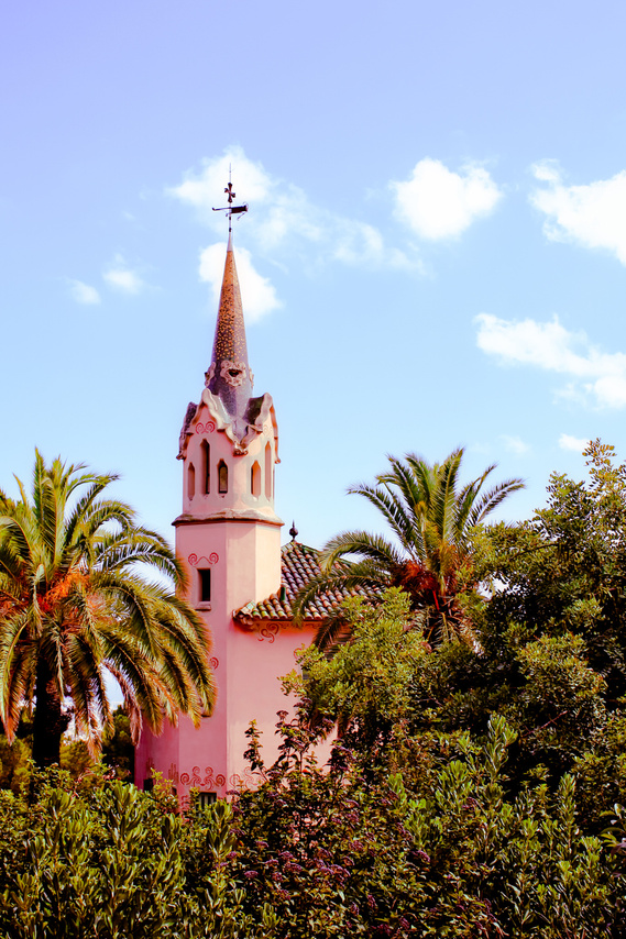 Iconic pink washed spanish style architecture with a pointed tower holding a weather vane set in lush palm trees and dense tropical plants set in front of a summer blue cloudy sky at Park Guell in barcelona Spain