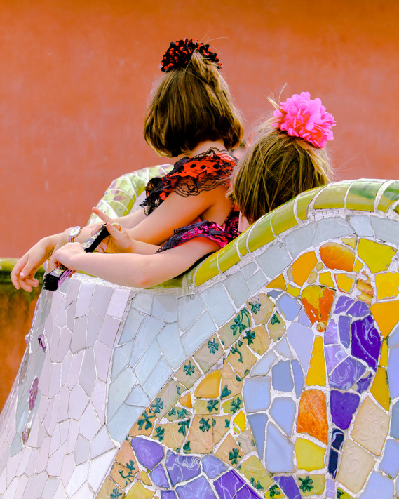 Portrait-oriented fine art photo print of two traditionally dressed flamenco girls in red and black polka dot dresses, fans and pink hair decorations leaning against the beautiful tile architecture by Antoni Gaudi of Park Guell in Barcelona
