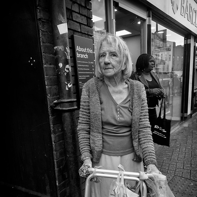 A woman with her walking frame standing outside shops in Tooting South London black-and-white photography