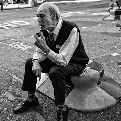 'Man sitting on a bollard smoking his pipe, Victoria train stationBlack & White London Street Photography'