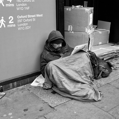 Rough sleeper sitting outside shop reading newspaper London black-and-white photography