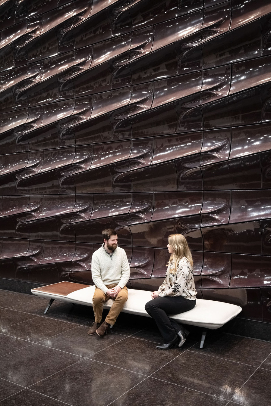 A couple sitting on a modern bench in front of a terracotta wall.