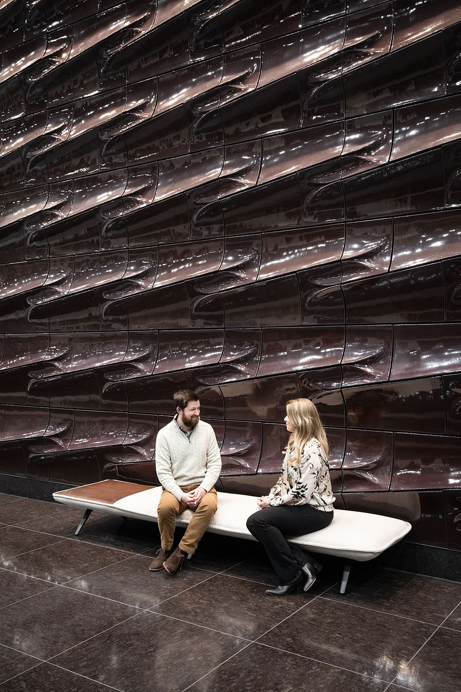 A couple sitting on a modern bench in front of a terracotta wall.