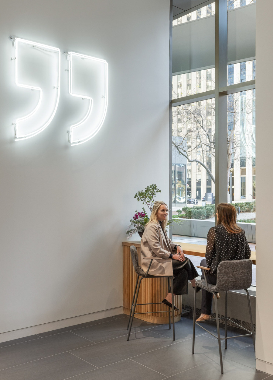 Two female coworkers sitting a bar in front of a large window with quotations light art on the wall.