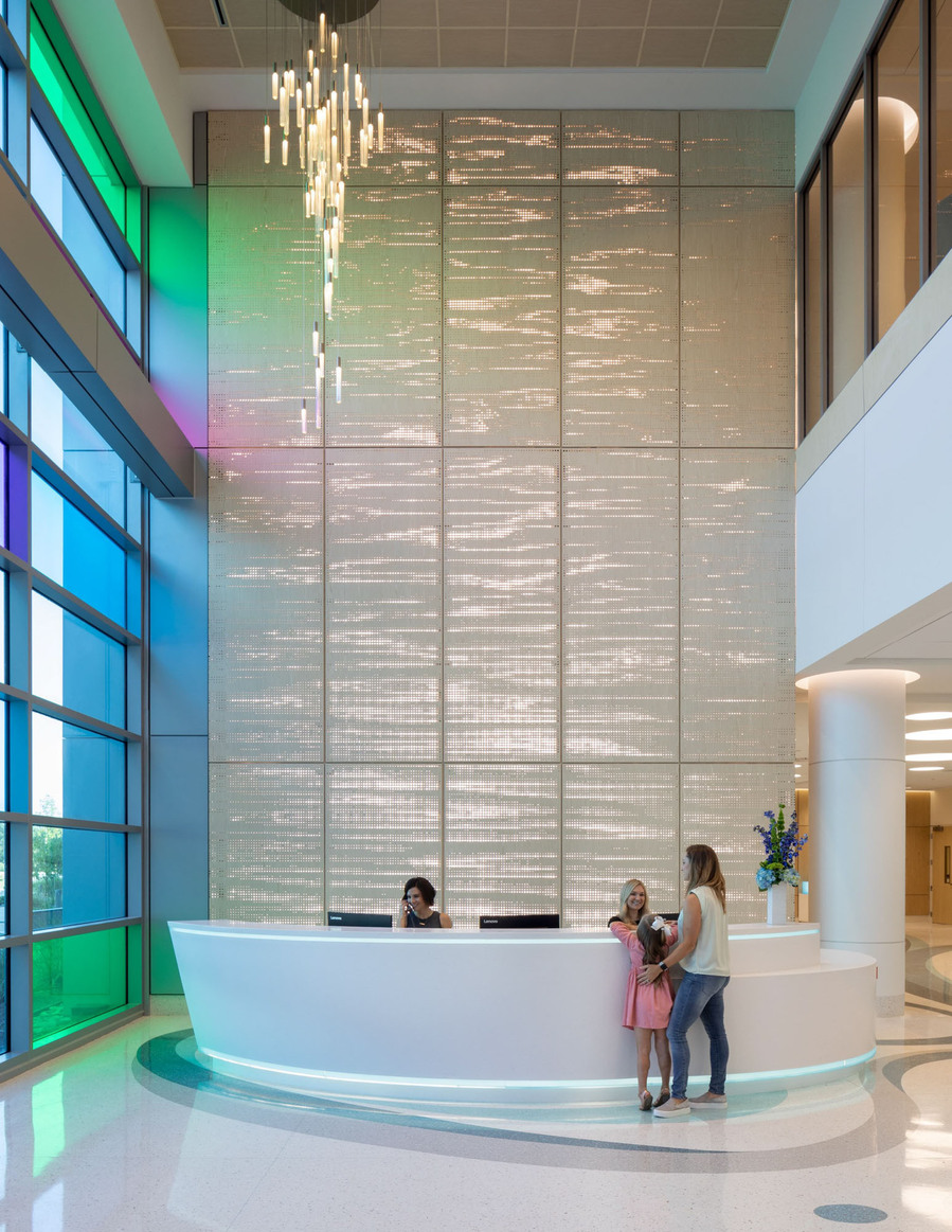 A large healthcare lobby with large windows, a chandelier and a women with her two daughters checking in.