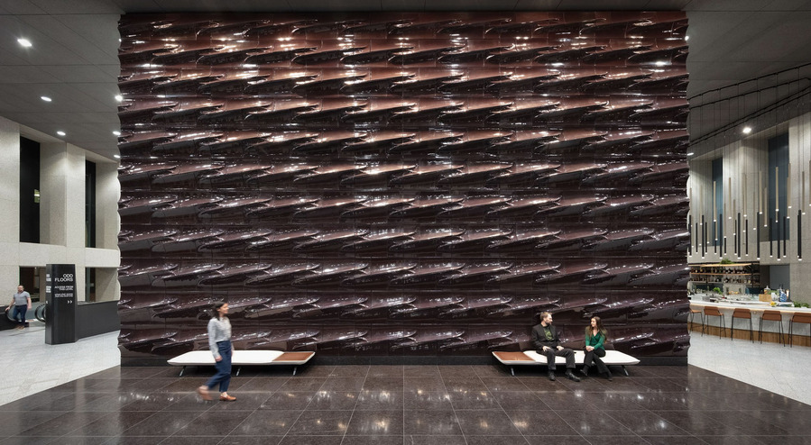 Large interior lobby with a massive terracotta wall with people sitting on a modern bench and a woman walking in motion.