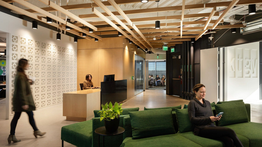 A modern office lobby with a woman sitting on a green sofa checking her cell phone. A woman at the front desk on her computer. A group meeting in a meeting room in the background.