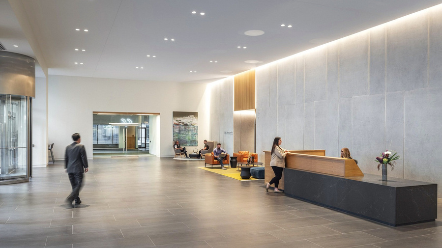 A modern office building lobby with a man walking in and a woman at the front desk speaking with a greeter. People sitting in the modern furniture in the background.