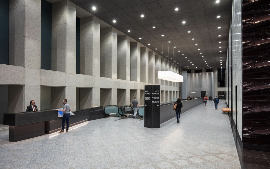 A massive corridor with a man speaking with the front desk, a man coming up the escalator and 3 people walking down the corridor.
