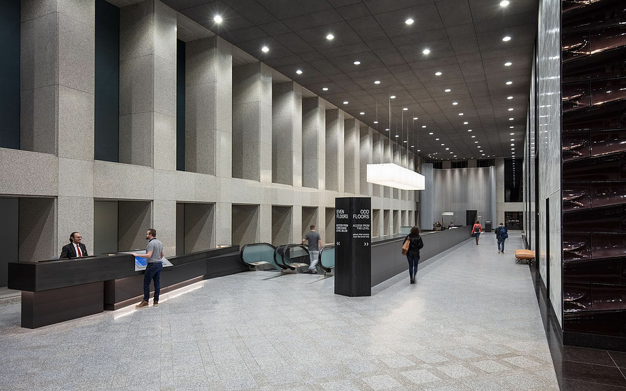 A massive corridor with a man speaking with the front desk, a man coming up the escalator and 3 people walking down the corridor.