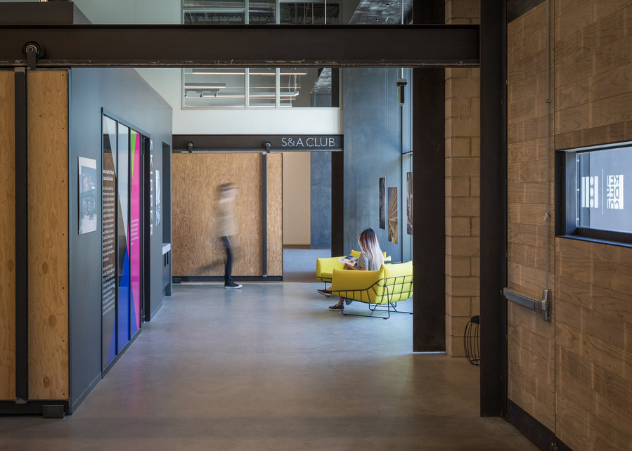 A modern office waiting area with large wood walls a women sitting in a yellow chair reading and a man in motion walking.