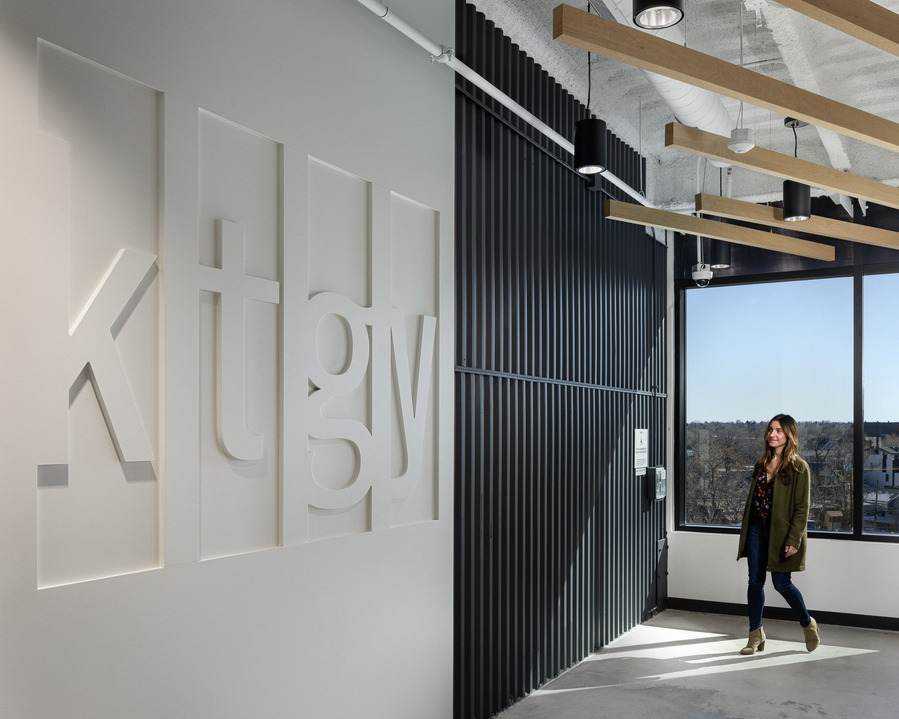 A women walking into an office entrance with KTGY on the wall, modern wood piece for the ceiling and a bright window with a view.