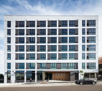 A modern white building with lots of glass, a wooden monument wall, a man walking down the sidewalk and Tesla parked out front. 