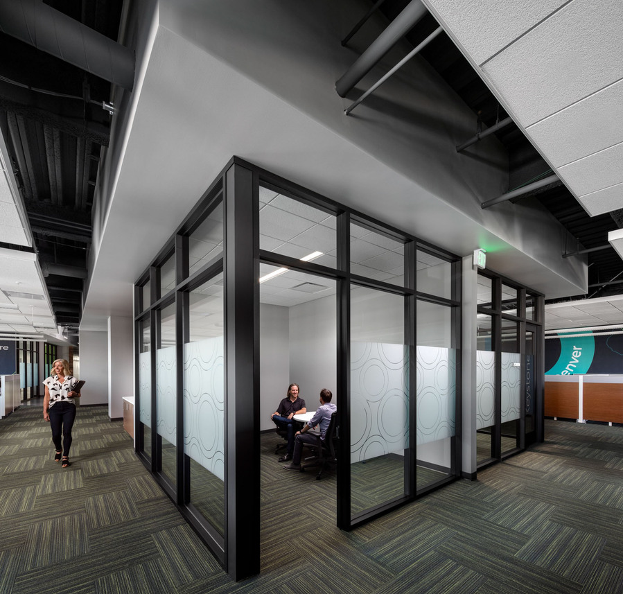 A modern office with woman walking down the hall and two men in a meeting inside the office.