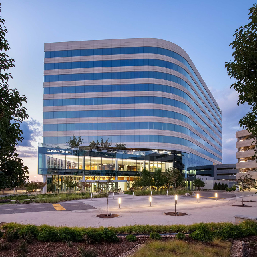 Office building with lots of glass framed by trees at dusk with interior lighting illuminating the foreground.