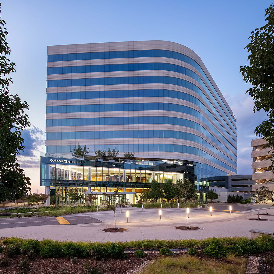 Office building with lots of glass framed by trees at dusk with interior lighting illuminating the foreground.