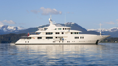 A luxury mega yacht during a bright blue day on the ocean waters of Alaska with a dramatic mountain scene in the background.