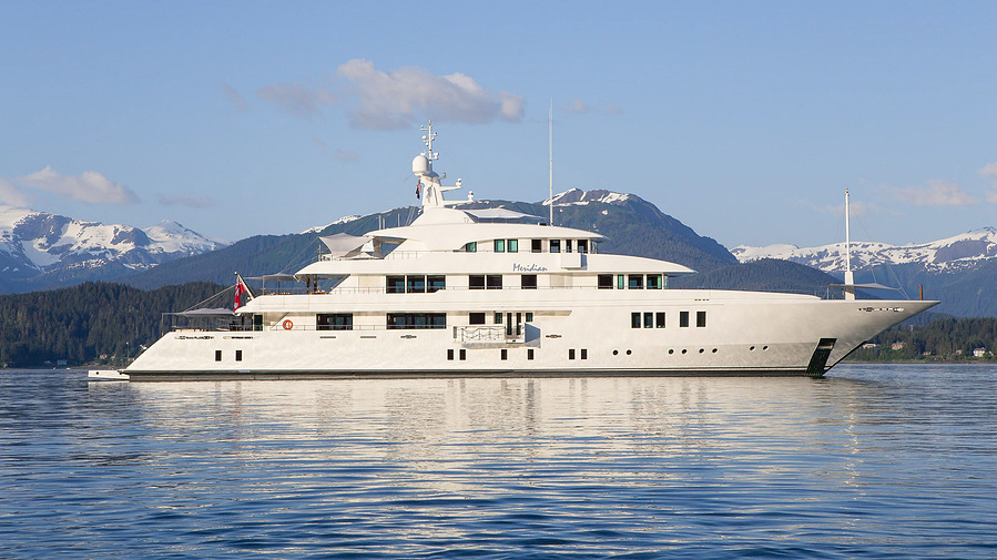 A luxury mega yacht during a bright blue day on the ocean waters of Alaska with a dramatic mountain scene in the background.