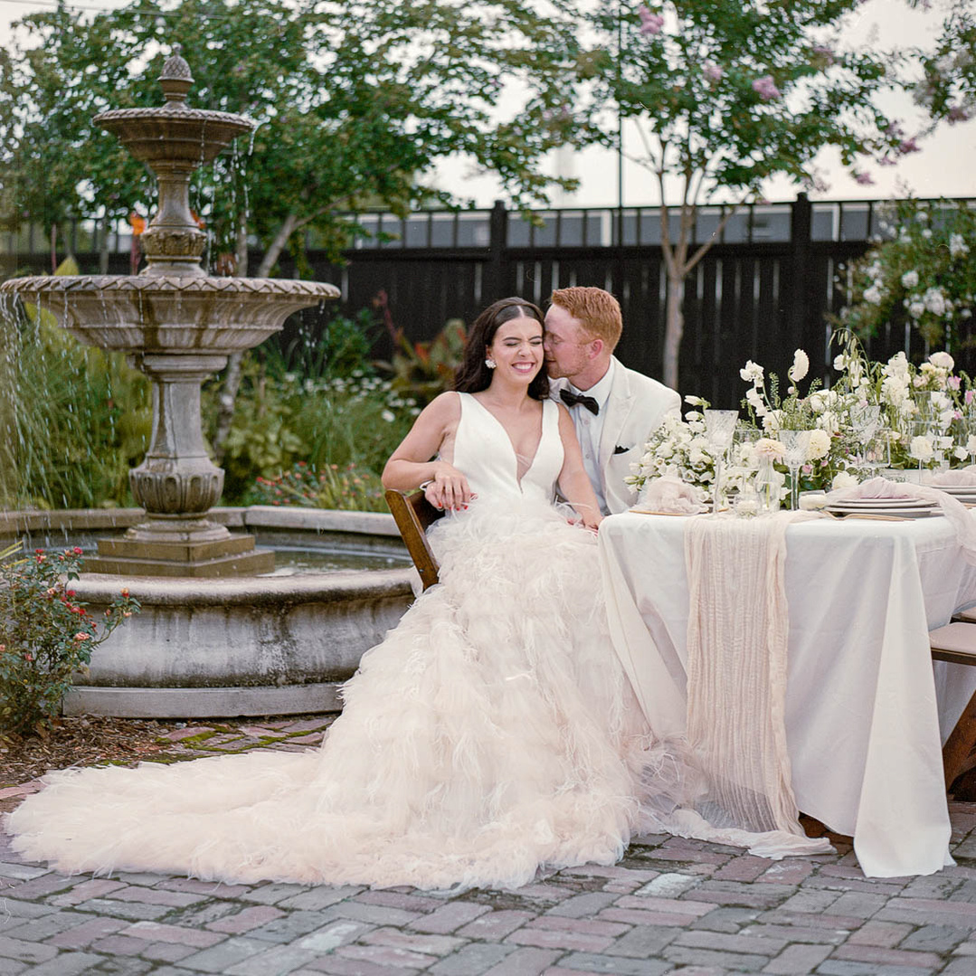 A couple dressed in wedding attire sits at an elegantly decorated outdoor table near a stone fountain, with the groom kissing the bride on the cheek.
