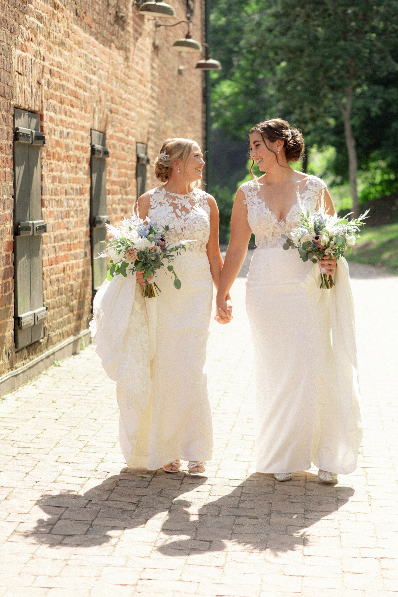 Two brides in white wedding dresses walk hand-in-hand on a sunlit brick pathway. Each holds a bouquet of flowers and smiles warmly at each other. Photographed in Roswell, GA