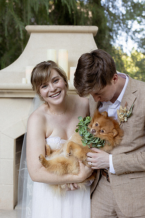 a candid photo of a bride and groom on their wedding day, laughing at the alter and holding a small dog wearing wedding attire