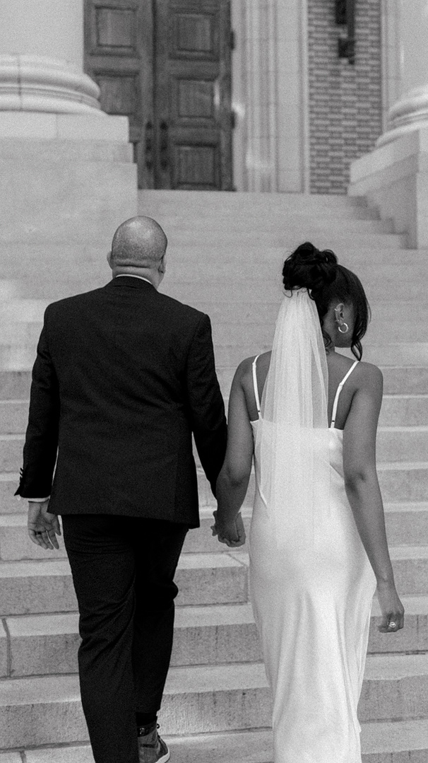 A couple, dressed in formal attire, walks up a set of stairs towards large wooden doors. The woman wears a white dress and veil, and the man is in a black suit. Atlanta Georgia Wedding Photoshoot