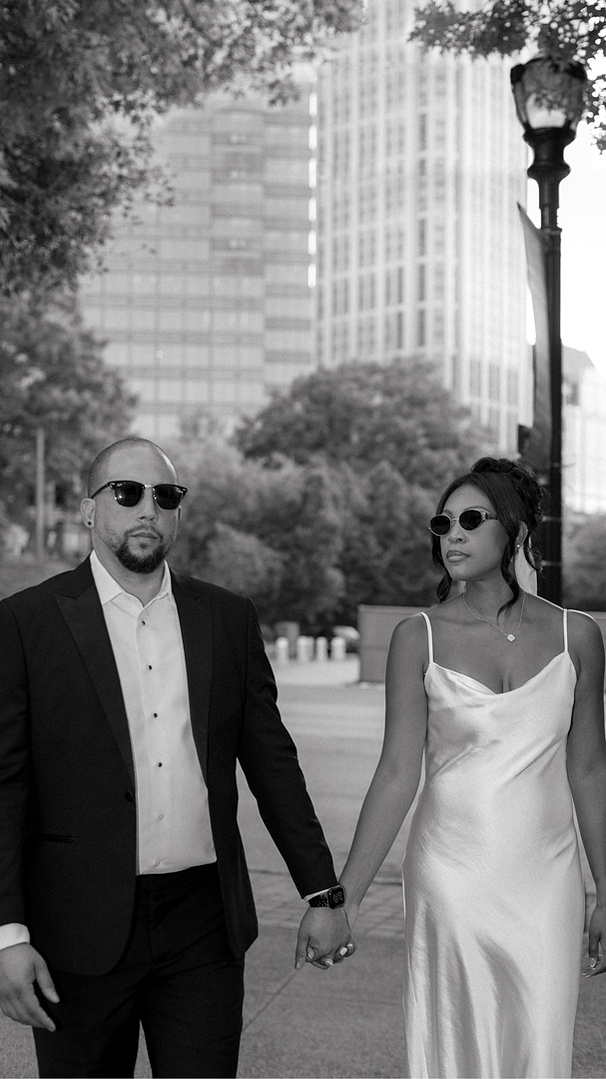 A couple holding hands while walking outdoors in a city, both wearing sunglasses and formal attire. Atlanta Wedding photography. Documentary Style
