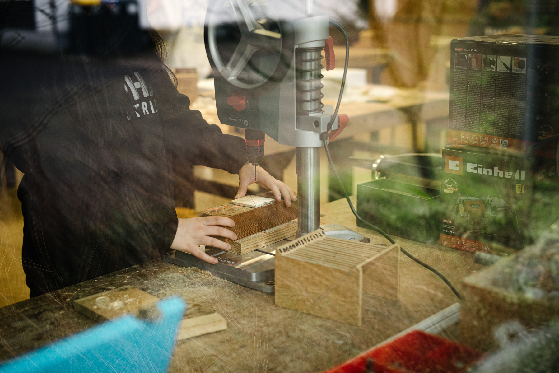 A student's hands, seen through a window, cut a piece of plywood in a shop classroom.