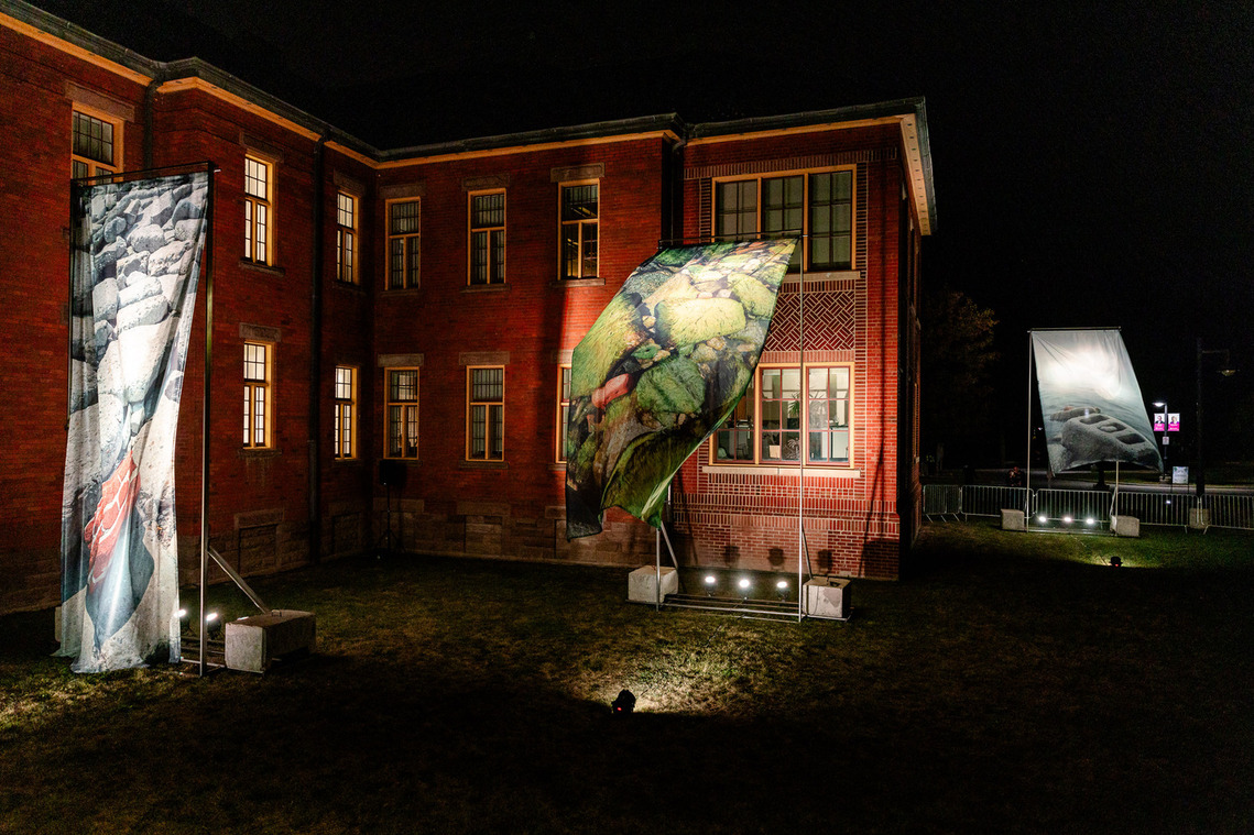 Digital images printed on sheer textiles, hang as oversized curtains in a courtyard, surrounded by red brick romanesque buildings from the former Lakeshore Psychiatric Hospital in Etobicoke.