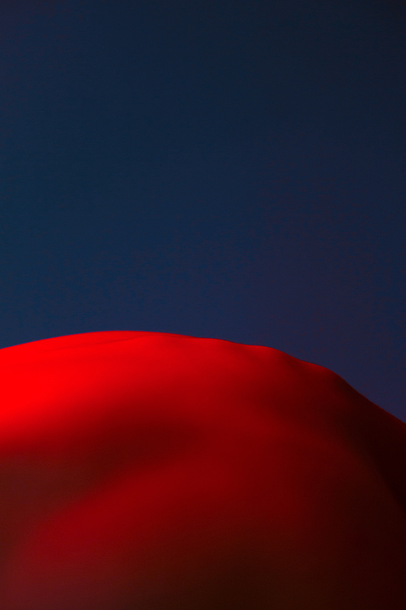 Man body in nude in red color and there is deep navy background it looks like a surreal landscape of a desert