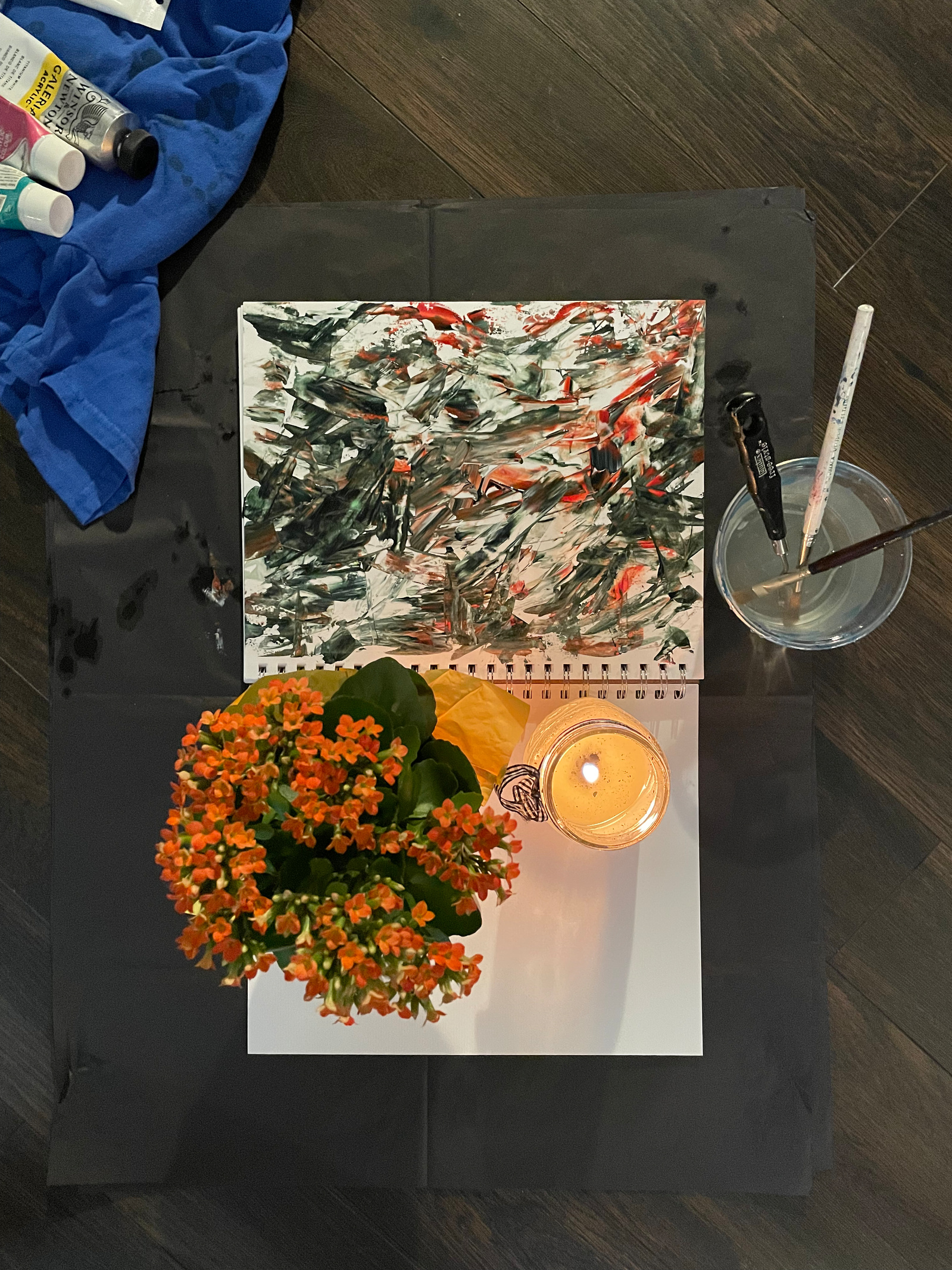 A painting with a candle, a plant with orange flowers and a water bowl with brushes on the side, ready for the camera.