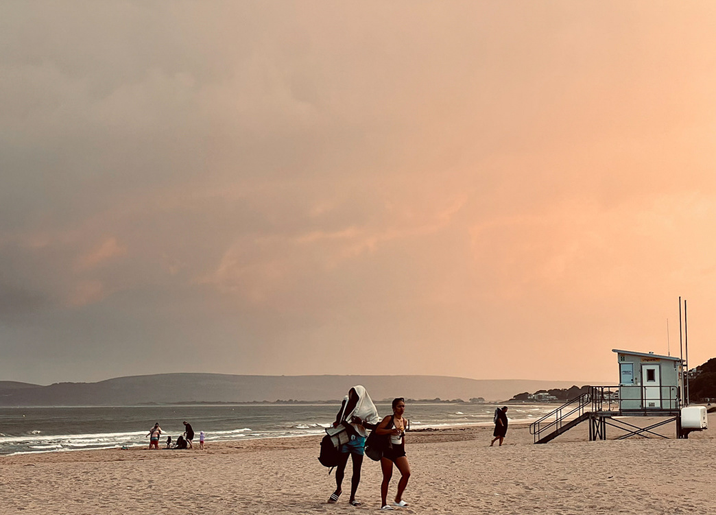 Two people leave the beach, one with a towel on their head against a sunset backdrop