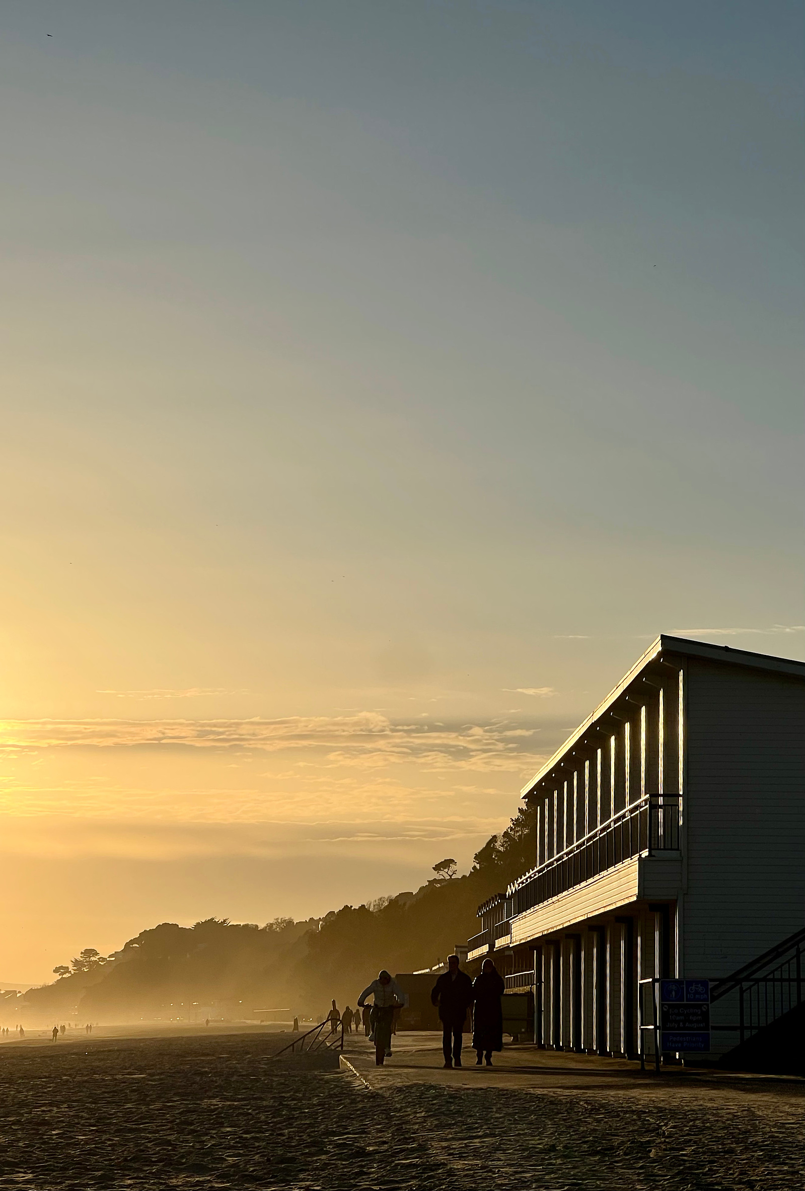 The setting sun hits the panels of a building; people can also be seen illuminated and walking along the promenade