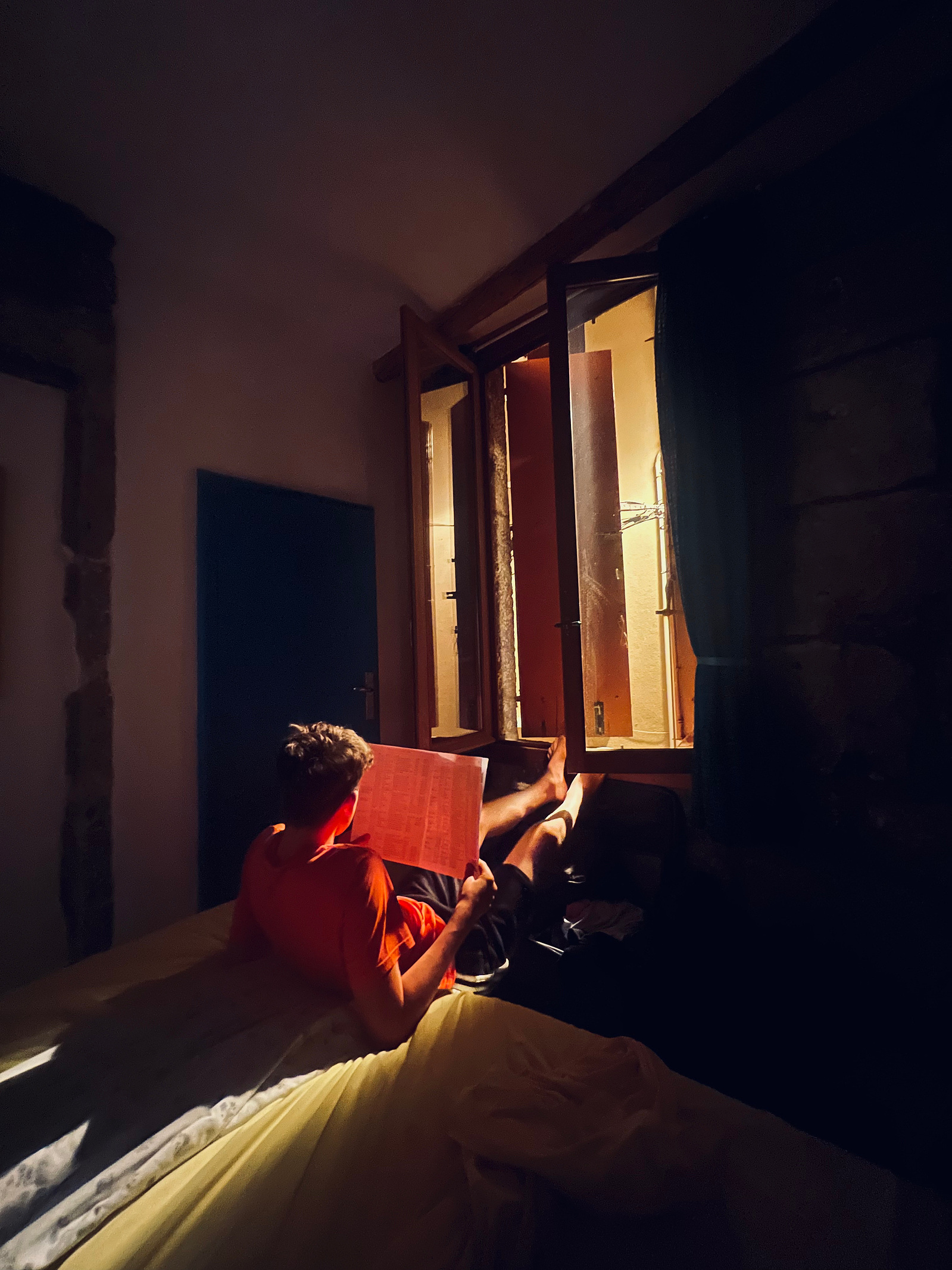 Light streams through a shutter casting a boy reading a book with light (Agde, France)
