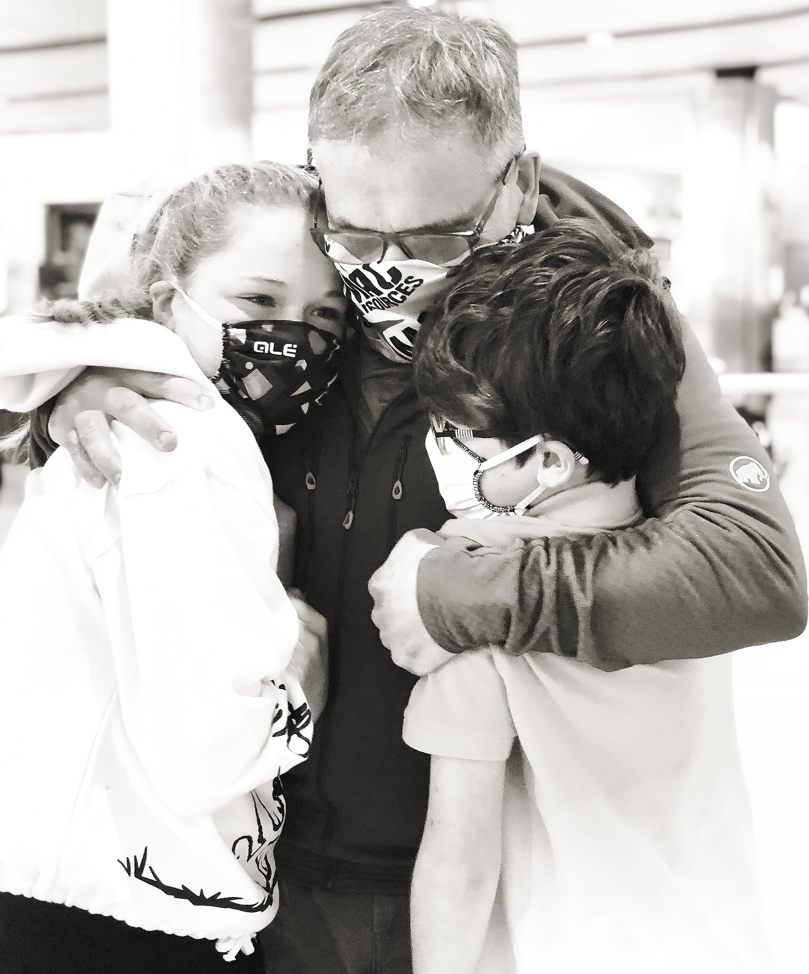A dad hugs his two children in arrivals at the airport. They are all wearing masks due to the restrictions in place due to Covid.