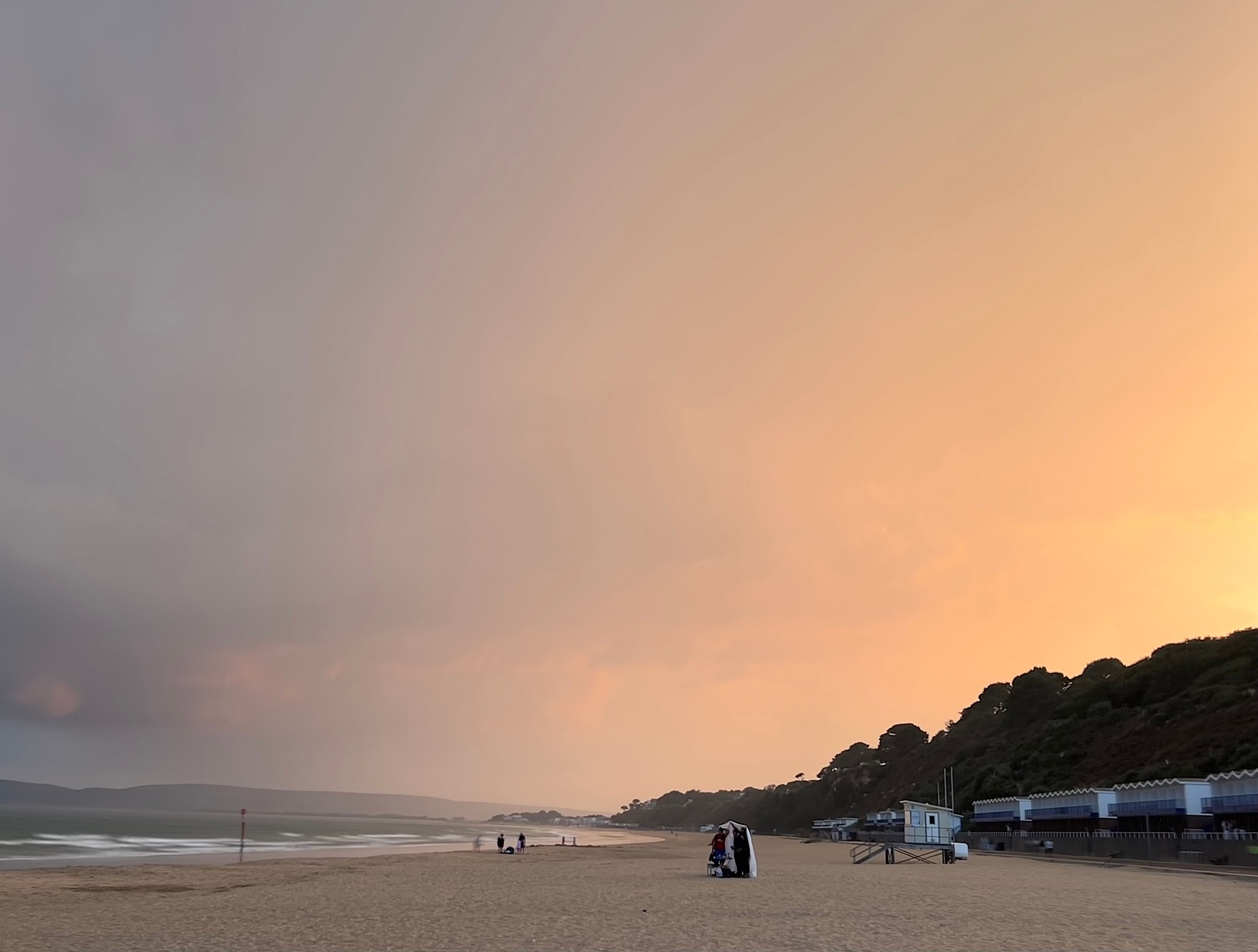 Orange light fills half of the frame balanced against some dark, stormy clouds moving on. Small figures contrast with the scale of the sky and the beach and one couple can be seen huddled under a towel in the mid-ground