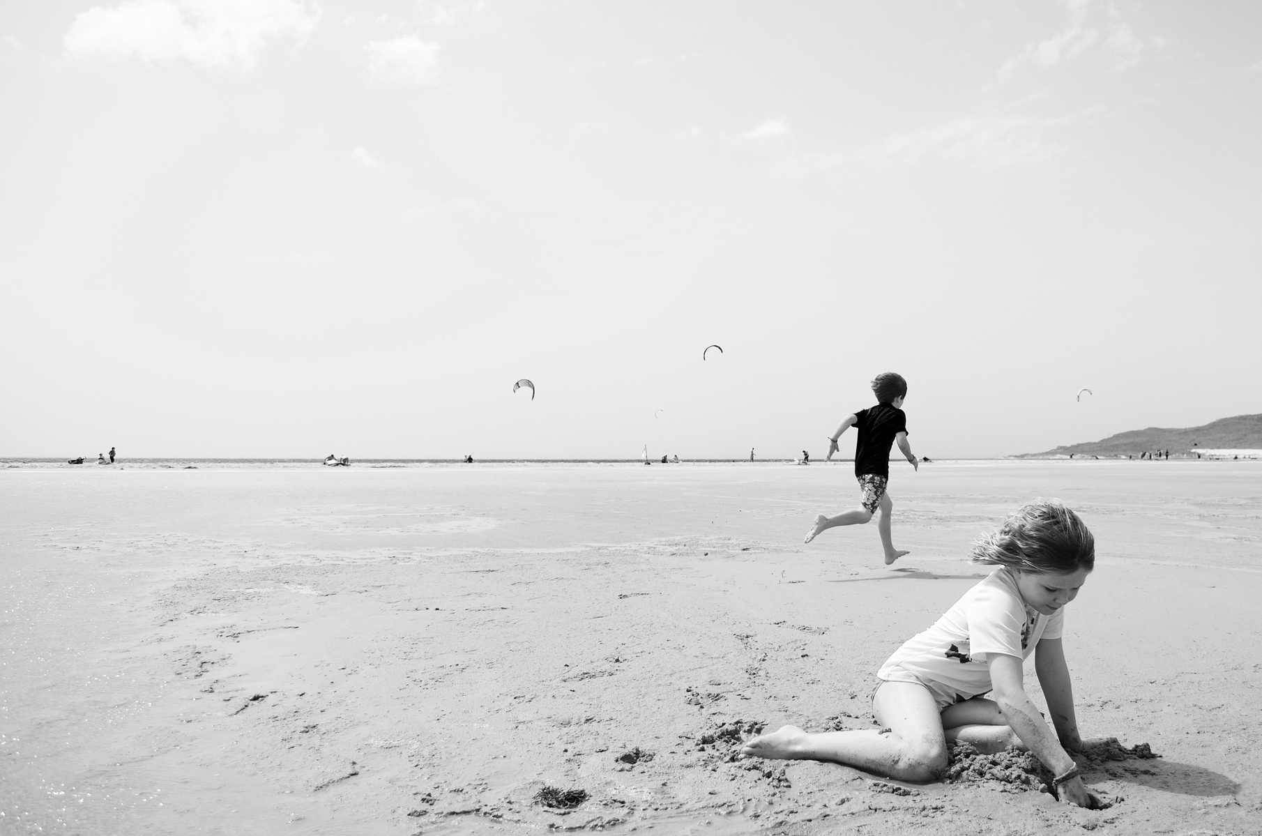 A young girl and her brother are playing on a beach in Tarifa, Spain with kite surfing in the background. The gils is playing with the sand the boys is running quickly behind her.