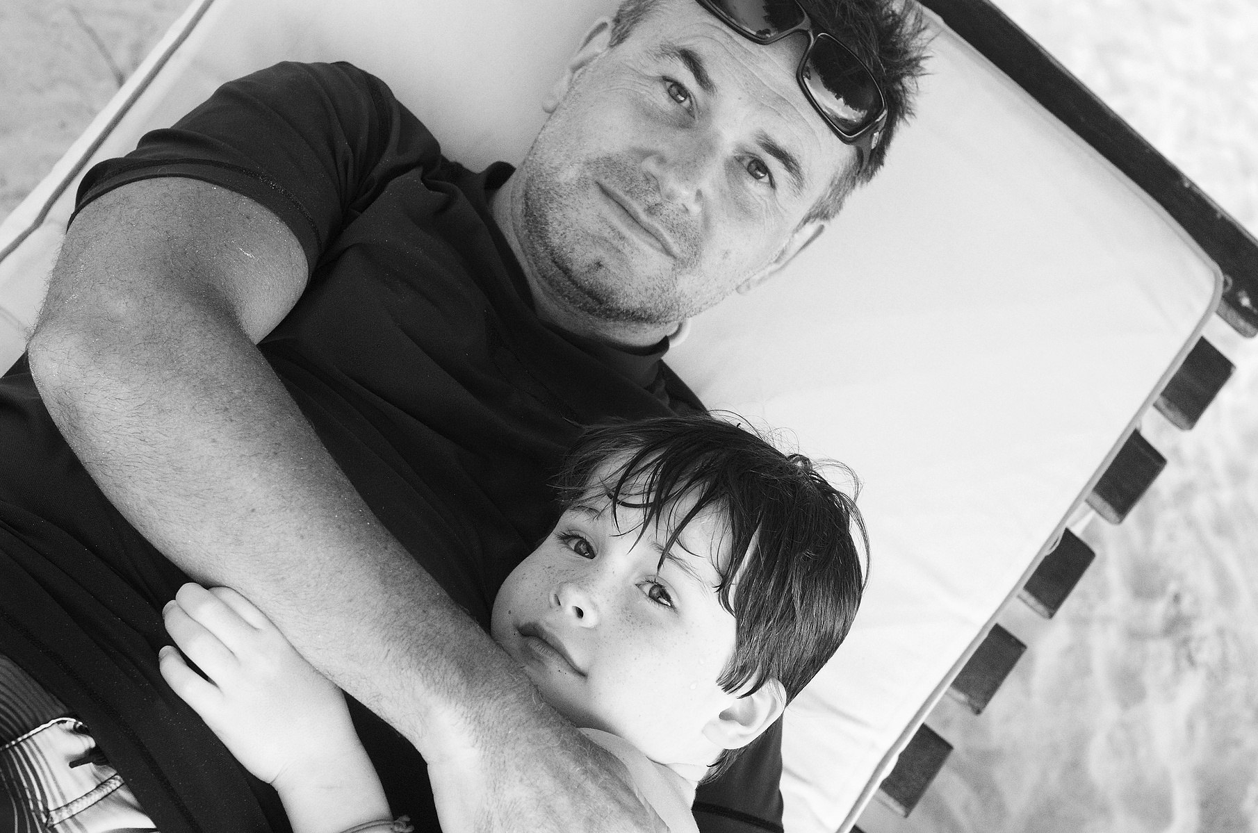 A father and his son lie on a sun bed looking directly up at the camera. The boy looks safe and secure, the dad looks peaceful and wraps his arms around his young son.